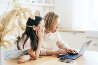 Mom and daughter at the table in the kitchen are looking at the laptop monitor.