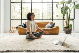 Young multiracial latina woman meditating at home with online video meditation lesson using laptop.